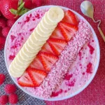 A colorful bowl filled with layers of creamy oats, bright pink raspberry yogurt, coconut flakes, and pistachio nuts.
