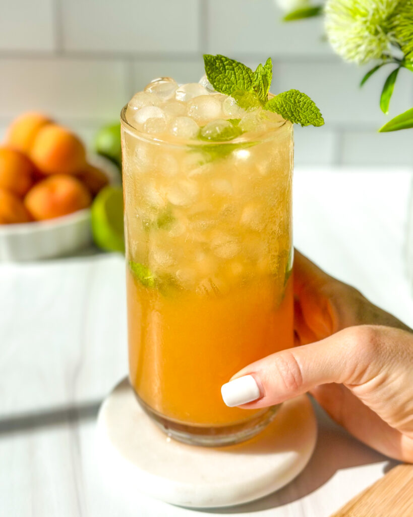 Hand holding a chilled apricot mojito garnished with mint on a sunny summer day