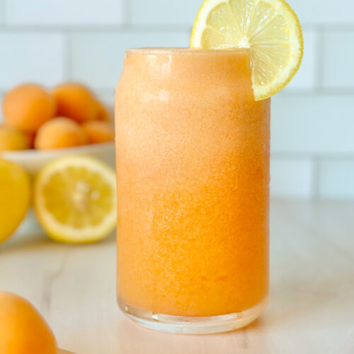 Two glasses of fresh apricot lemonade served chilled, styled with apricots and sliced lemon in the background for a bright summer drink scene.