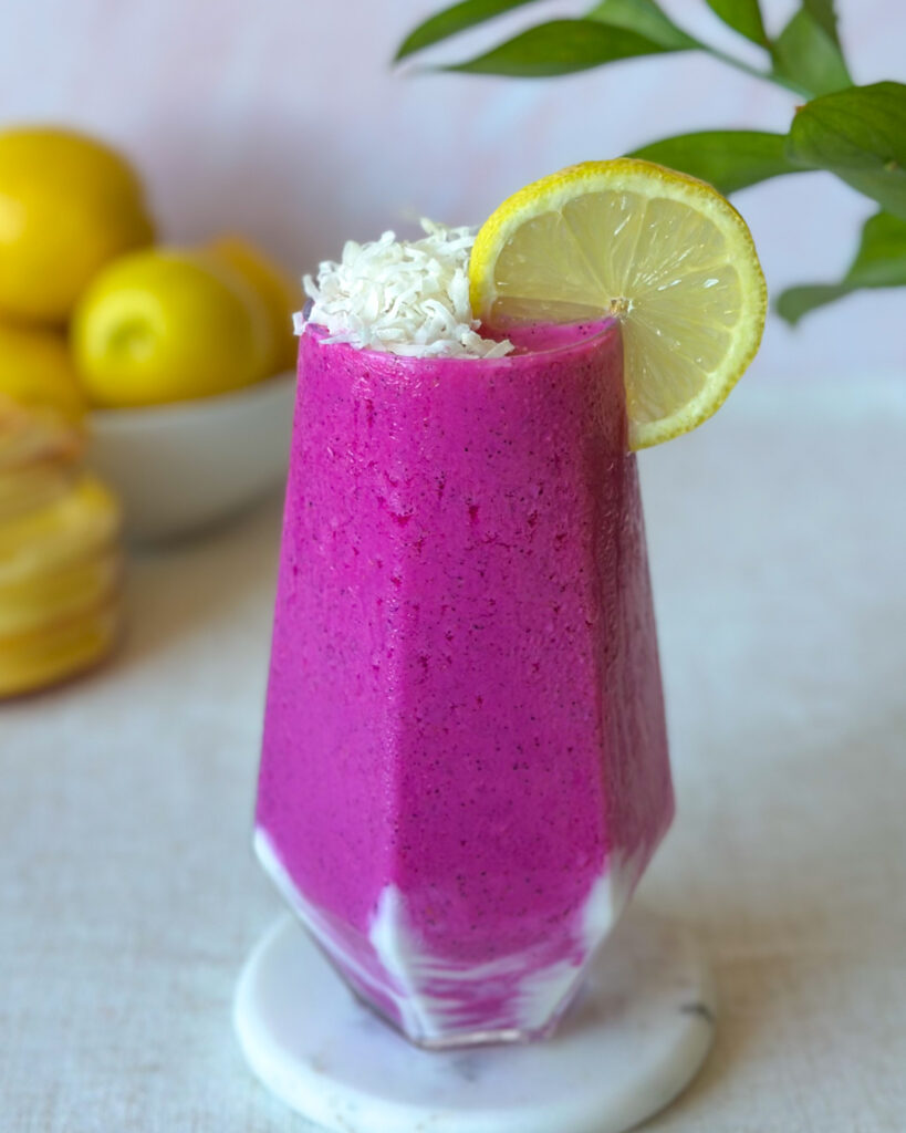 slightly overhead view of vibrant pink dragon fruit smoothie with lemons and tropical green leaves in the background