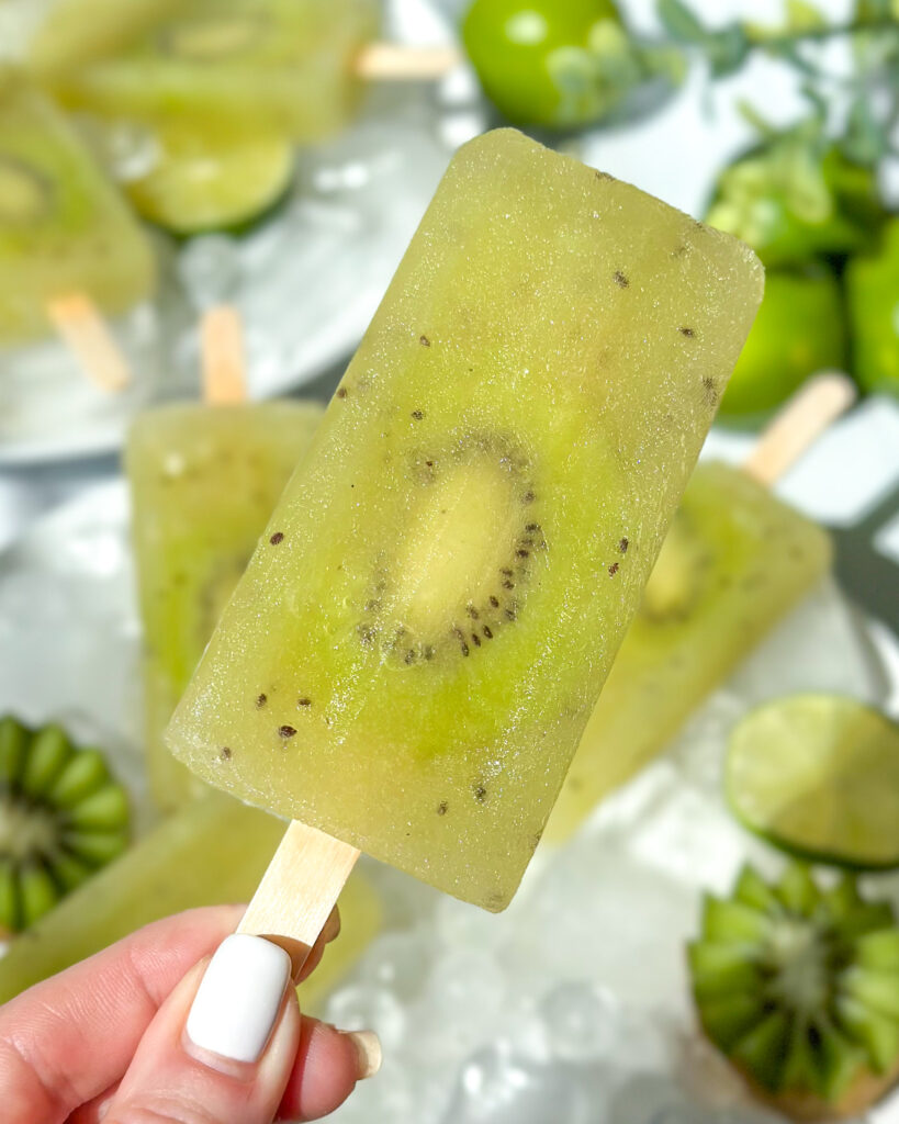 Close-up of a kiwi popsicle with pineapple and coconut water being held in the foreground with plates of popsicles softly blurred in the background.