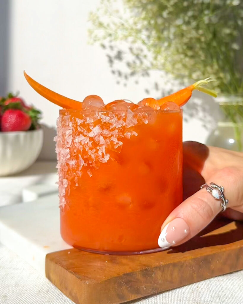 Hand holding a finished strawberry carrot margarita with a salted rim, carrot garnish, and ice cubes, styled with baby’s breath flowers and strawberries on a marble and wood serving board.