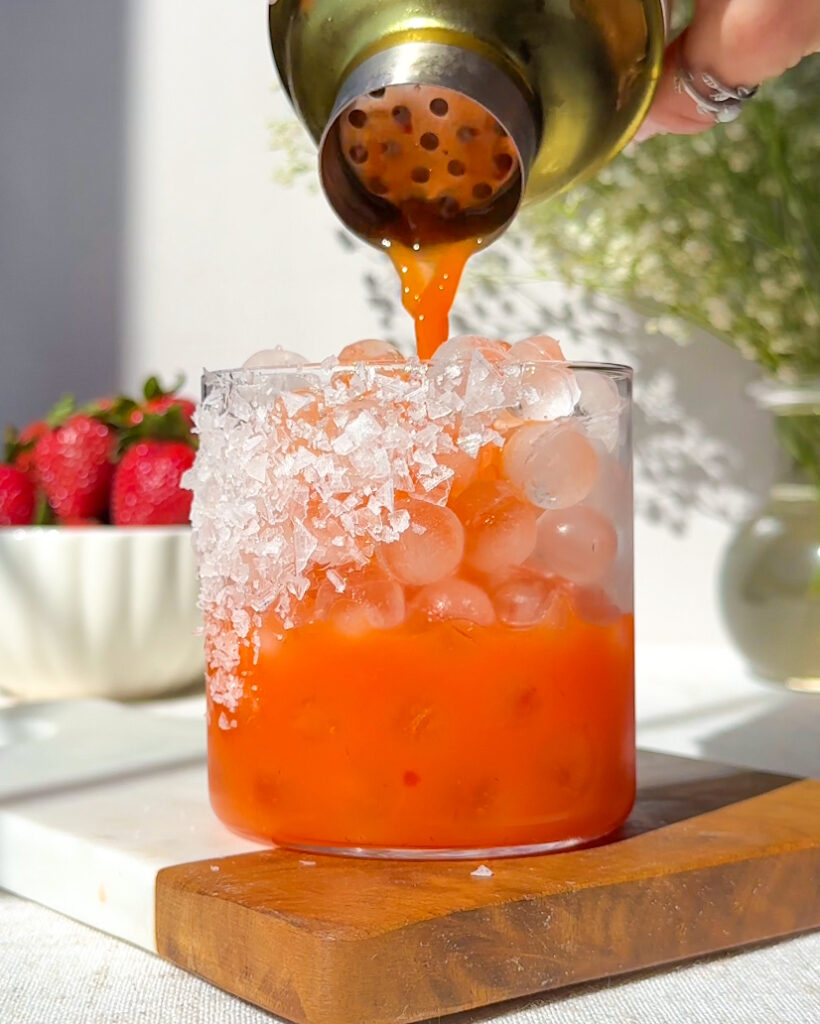 Action shot of strawberry carrot margarita being poured into a chilled glass filled with ice and a salted rim, with strawberries and baby’s breath styled in the background.