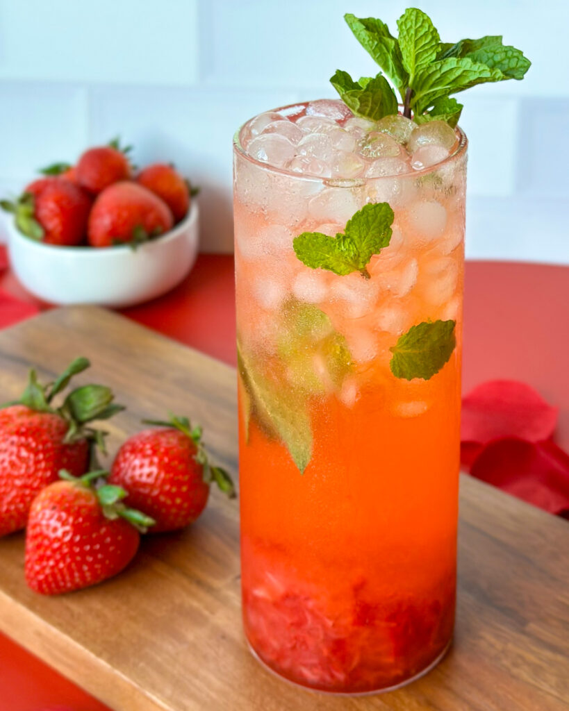 Strawberry mojito mocktail on a red surface with a wooden cutting board, strawberries, and red flowers in the background against a white tiled kitchen backdrop.
