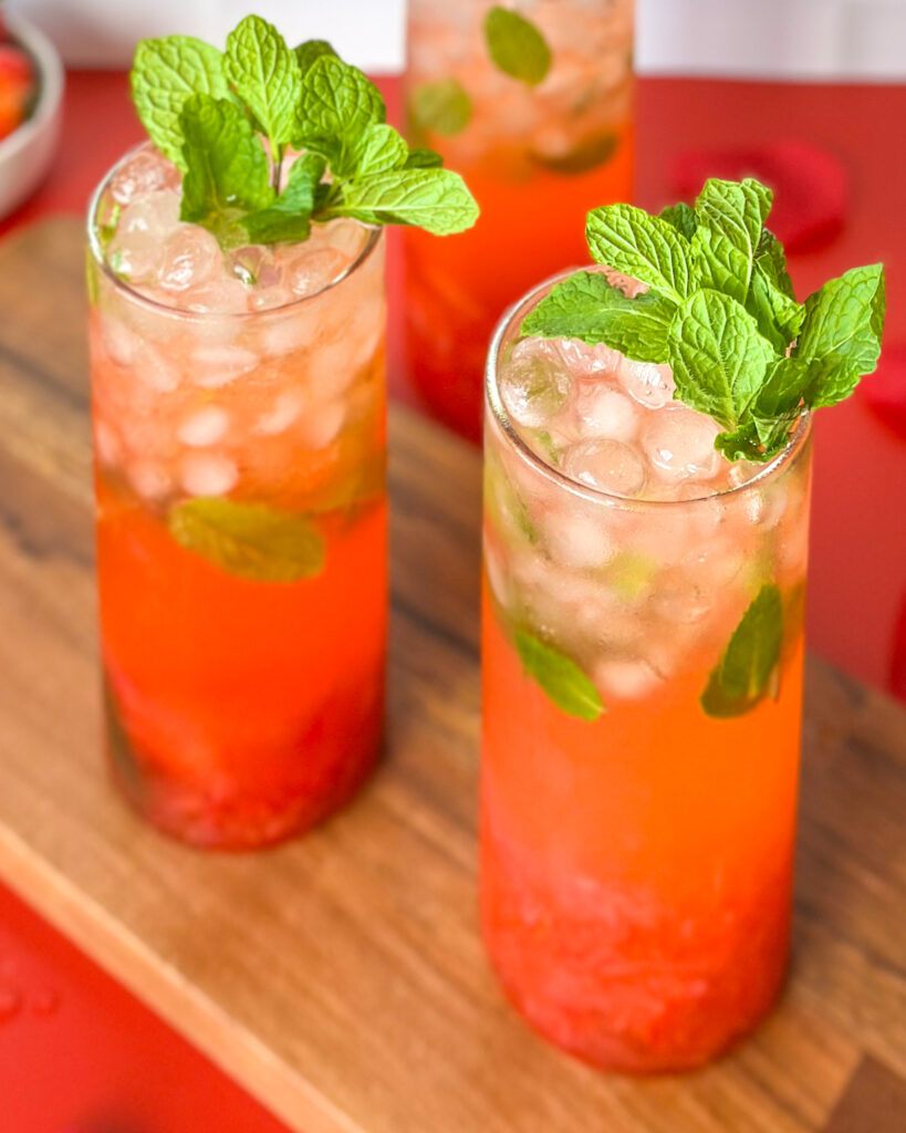 Two strawberry mojito mocktails served side by side in glasses, slightly angled front view on a red surface with a white tiled kitchen background.