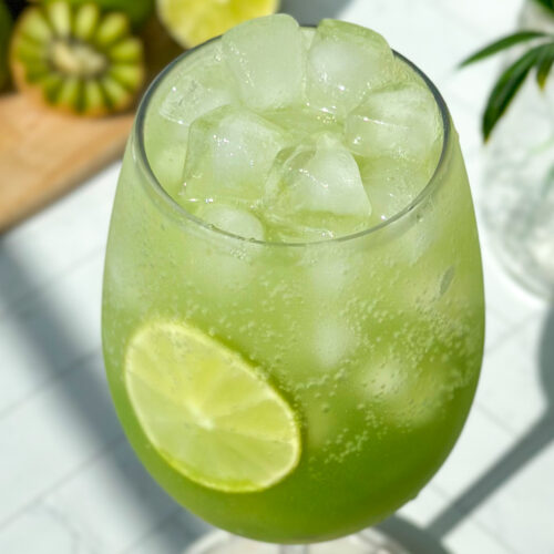 Extreme close-up overhead of a finished sparkling kiwi cucumber mocktail filled with ice, showing a dark green gradient at the bottom and a lime slice inside the glass, styled with kiwi, limes, florals, and a marble cutting board in the background.
