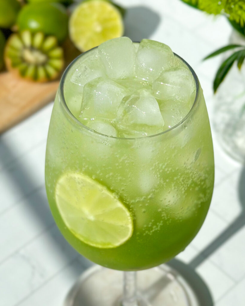 Extreme close-up overhead of a finished sparkling kiwi cucumber mocktail filled with ice, showing a dark green gradient at the bottom and a lime slice inside the glass, styled with kiwi, limes, florals, and a marble cutting board in the background.