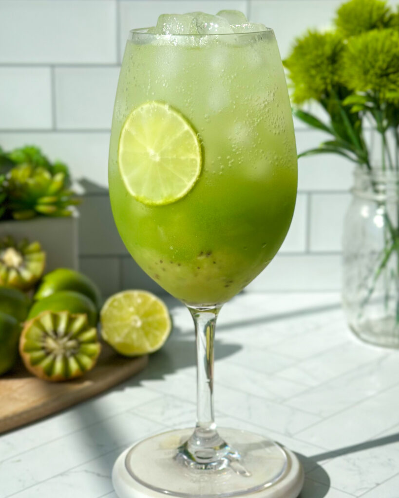 Close-up of a layered kiwi cucumber mocktail before stirring, showing distinct kiwi puree, cucumber juice, and sparkling soda layers in a glass on a marble surface with white tile backsplash, kiwi, lime, succulents, and green florals in the background.