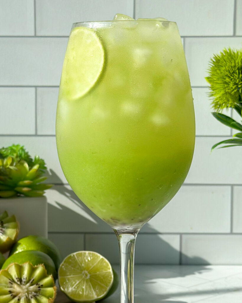 Extreme close-up of a fully stirred sparkling kiwi cucumber mocktail, now a vibrant light green color, with a lime slice floating inside the glass and styled against a white tile and fresh kiwi and lime background.