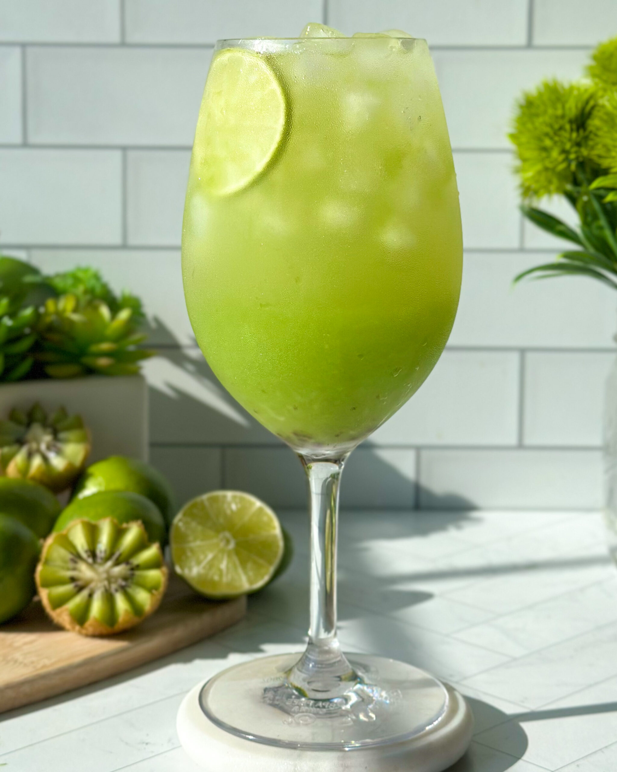 Front-facing, slightly pulled-back shot of a stirred sparkling kiwi cucumber mocktail with a vibrant green color and a lime slice floating at the top of the glass, styled with fresh kiwi, lime, and soft green background elements for a bright summer drink aesthetic.