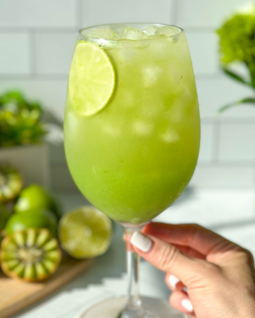 Portrait-style image of a hand holding a glass of sparkling kiwi cucumber mocktail, showing a softly blurred background with kiwi, lime, and green floral styling elements for a fresh summer drink aesthetic.
