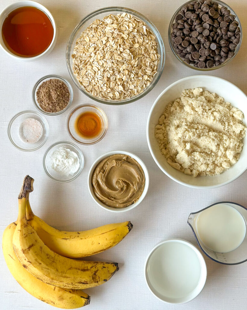 Flat lay of ingredients for banana oat chocolate chip bars including oats, almond flour, bananas, maple syrup, chocolate chips, and nut butter on a wooden surface.