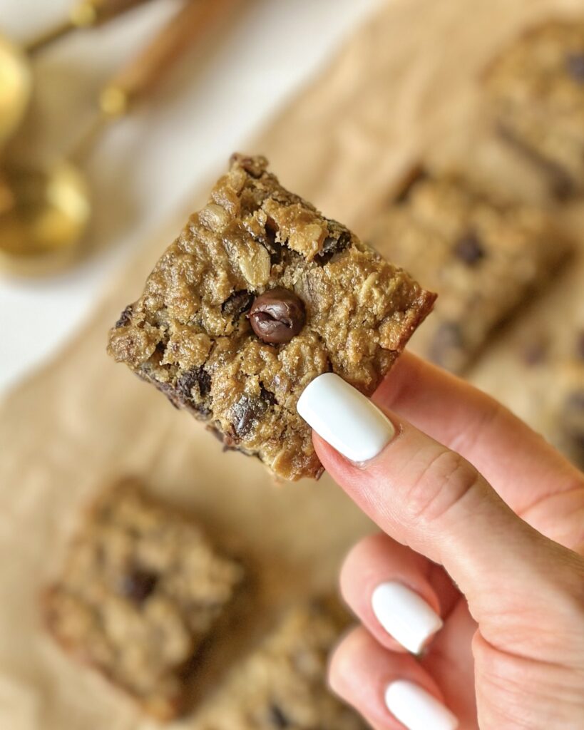 Extreme close-up of a soft banana oat chocolate chip bar held in hand with blurred background showing more oat bars behind.