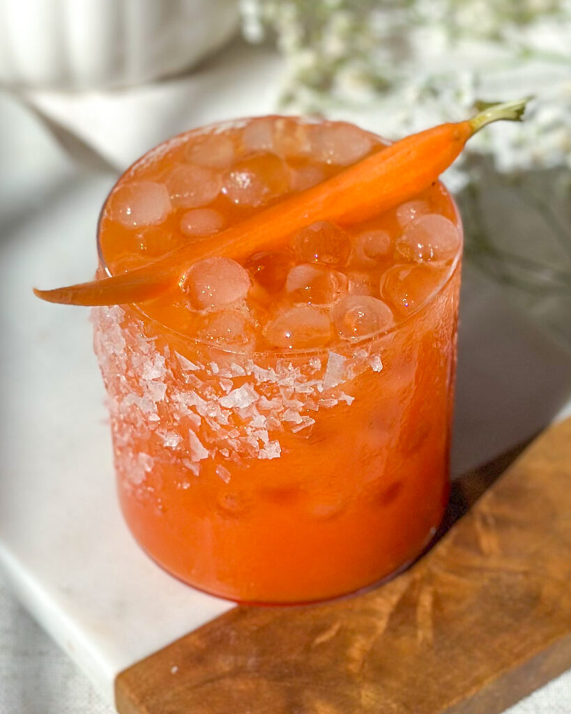 Angled front view of a strawberry carrot margarita showing the salted rim and carrot garnish, placed on a marble and wood serving board with baby’s breath flowers in the background.