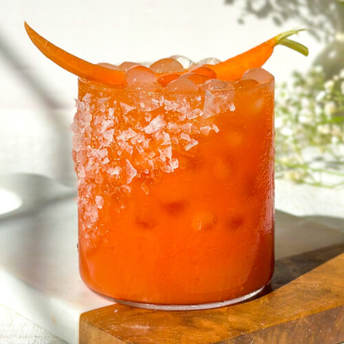 Front view hero image of a strawberry carrot margarita with a salted rim, vibrant orange color, and carrot garnish, styled on a marble and wood serving board with soft floral background.