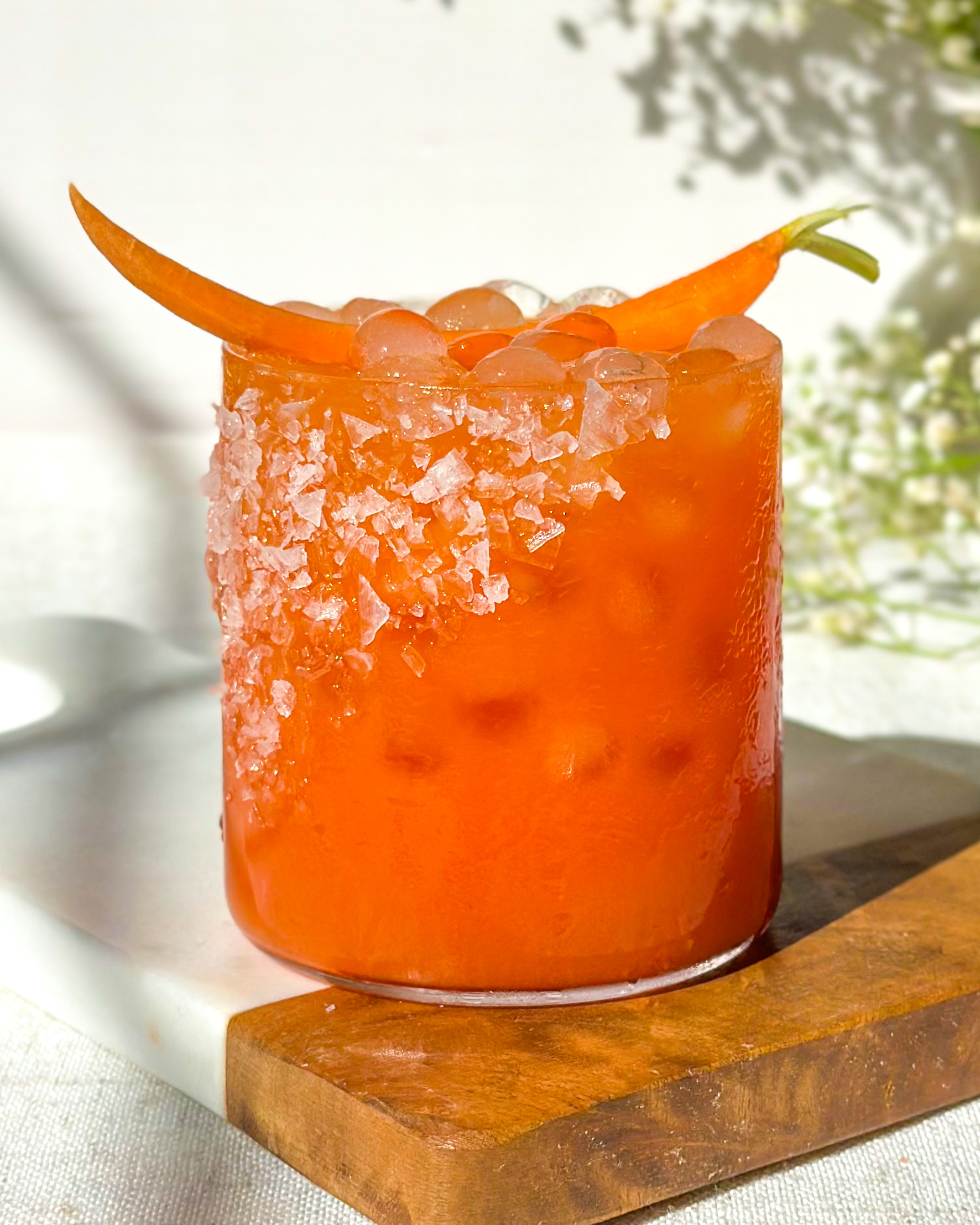 Front view hero image of a strawberry carrot margarita with a salted rim, vibrant orange color, and carrot garnish, styled on a marble and wood serving board with soft floral background.