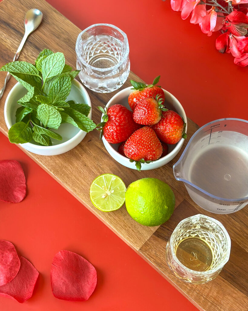 Flat lay of strawberry mojito mocktail ingredients including fresh strawberries, limes, and coconut water in bowls on a red surface with a wooden cutting board
