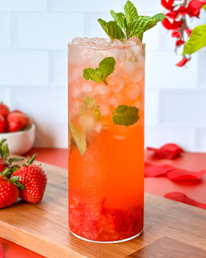 Front-facing strawberry mojito mocktail served in a glass on a red surface with strawberries in the background, a wooden cutting board, and a white tiled kitchen backdrop.