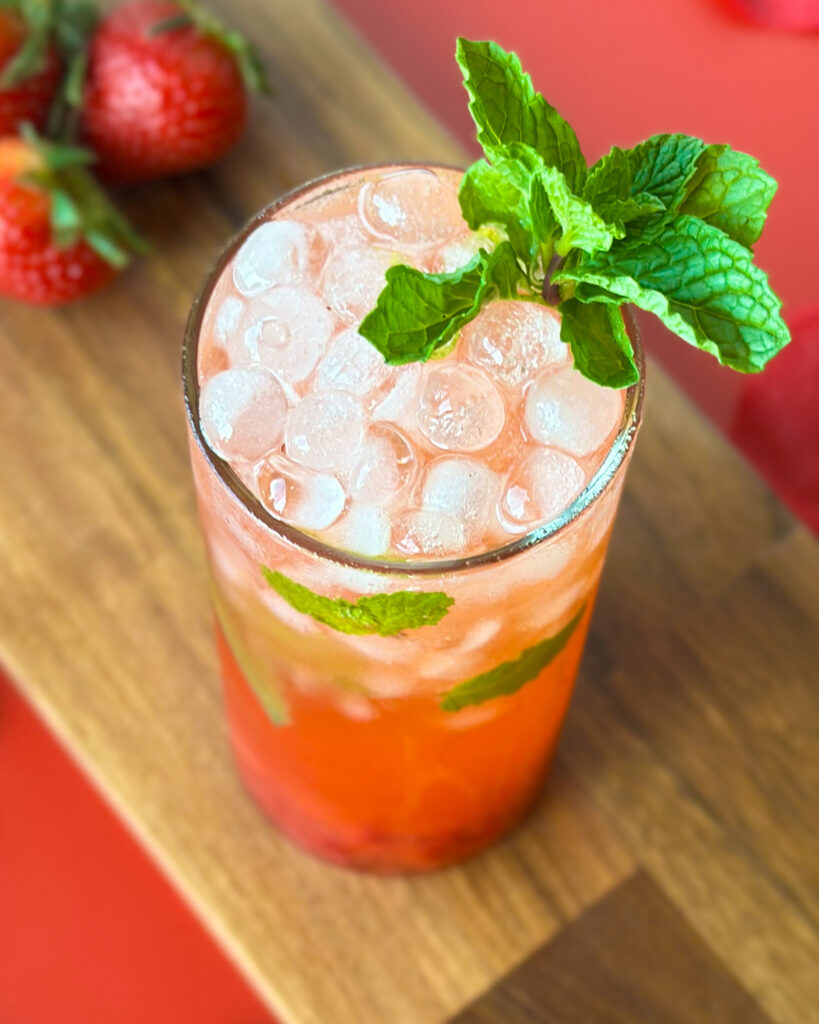Overhead view of a strawberry mojito mocktail with fresh mint and ice cubes, served on a red surface with strawberries and a wooden cutting board in the corner.