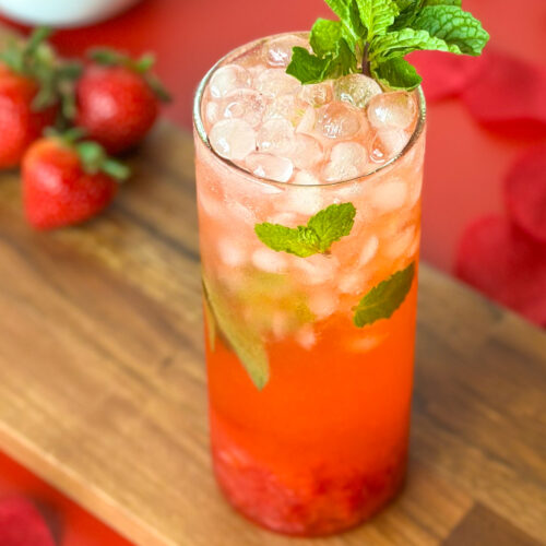 A refreshing strawberry mojito mocktail served in a glass, photographed at a slight angle with fresh strawberries, rose petals, and a wooden cutting board on a red surface in the background.