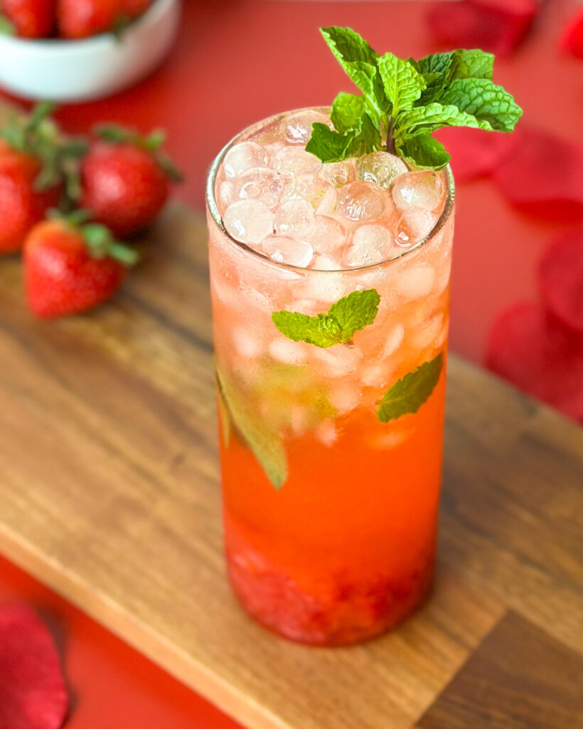 A refreshing strawberry mojito mocktail served in a glass, photographed at a slight angle with fresh strawberries, rose petals, and a wooden cutting board on a red surface in the background.