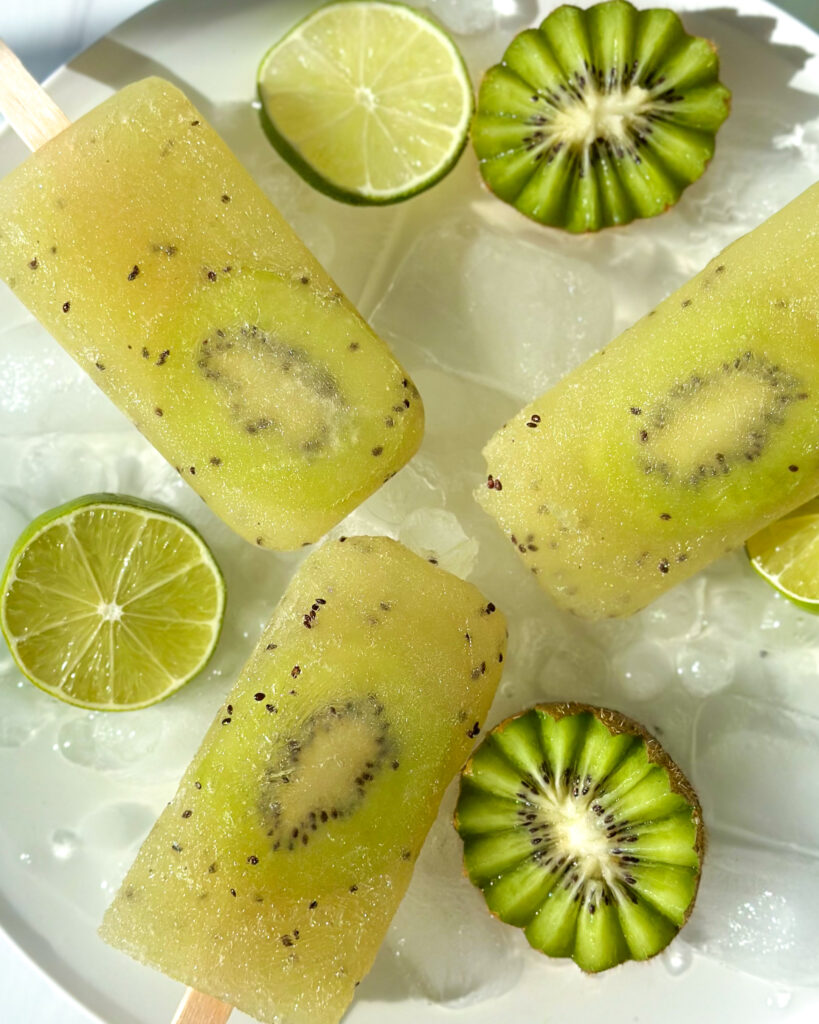 Overhead close-up of three kiwi popsicles on a bed of ice with sliced kiwi flowers and lime garnish