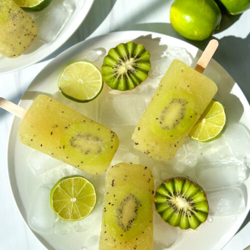 Overhead styled food scene featuring kiwi popsicles on ice with lime slices and garnishes, with additional plates arranged nearby