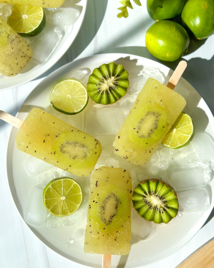Overhead styled food scene featuring kiwi popsicles on ice with lime slices and garnishes, with additional plates arranged nearby