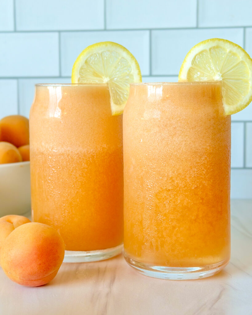 Two glasses of fresh apricot lemonade served chilled, styled with apricots and sliced lemon in the background for a bright summer drink scene.