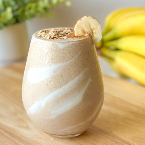 Front-facing hero image of a banana bread smoothie with a yogurt swirl inside the glass, topped with oats, cinnamon, and banana slice, styled with bananas in the background.