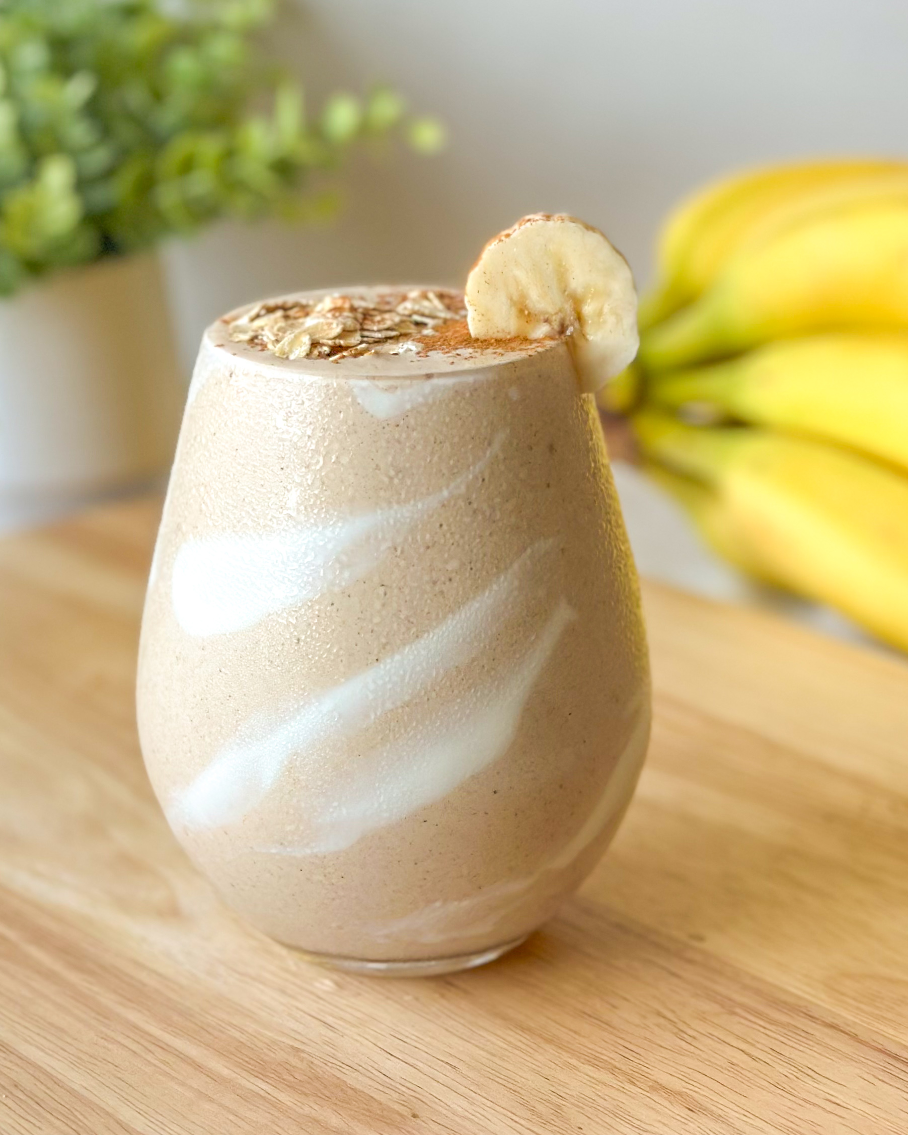 Front-facing hero image of a banana bread smoothie with a yogurt swirl inside the glass, topped with oats, cinnamon, and banana slice, styled with bananas in the background.