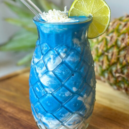 close up front on view of pineapple coconut blue spirulina smoothie with shredded coconut, lime wheel and a whole pineapple in the background