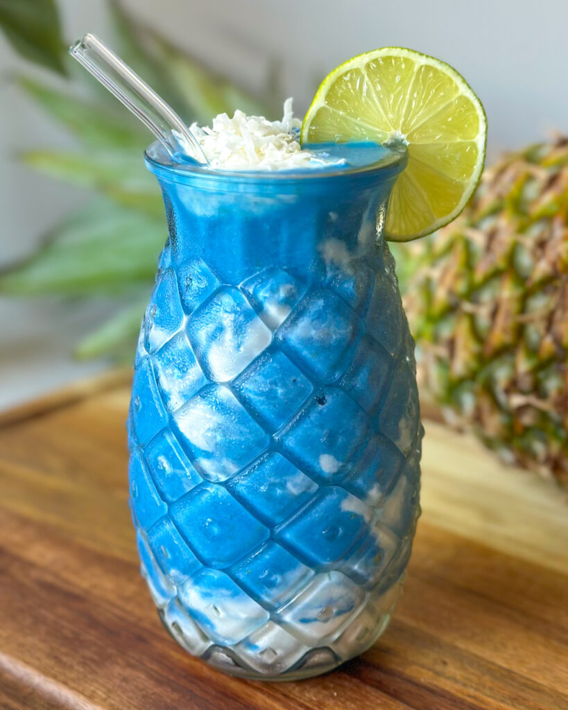 close up front on view of pineapple coconut blue spirulina smoothie with shredded coconut, lime wheel and a whole pineapple in the background