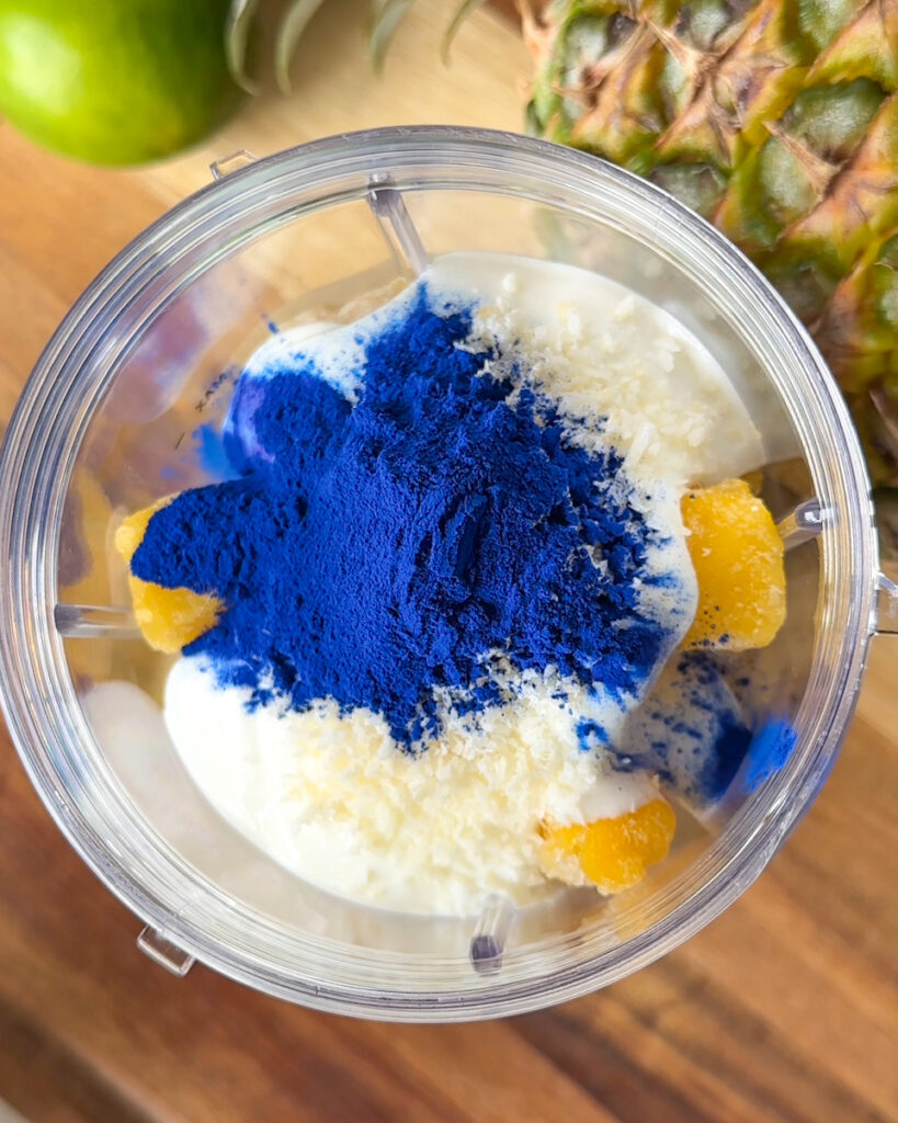 close up overhead view of frozen pineapple banana, grapes, mango yogurt coconut and blue spirulina in blender cup