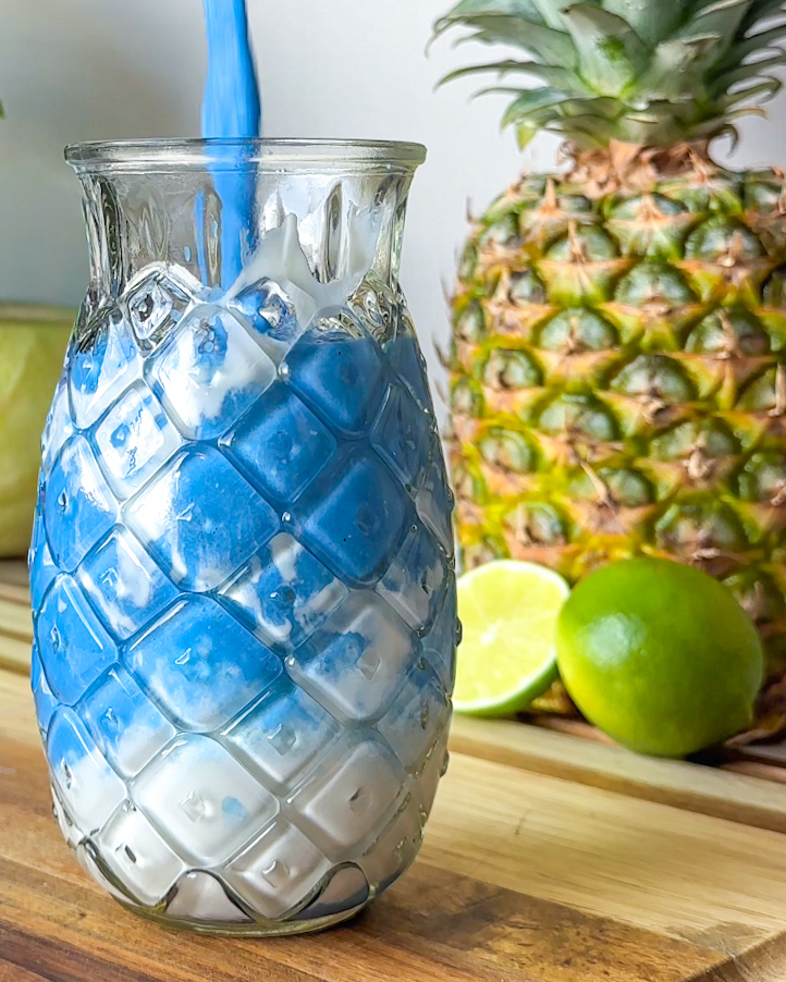 pouring pineapple coconut blue spirulina smoothie into a chilled pineapple shaped glass with yogurt swiped inside