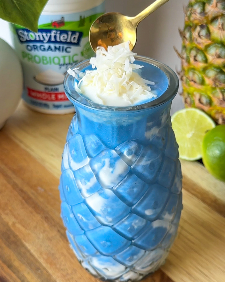 spooning shredded coconut on top of a finished pineapple coconut blue spirulina smoothie served in a pineapple shaped glass