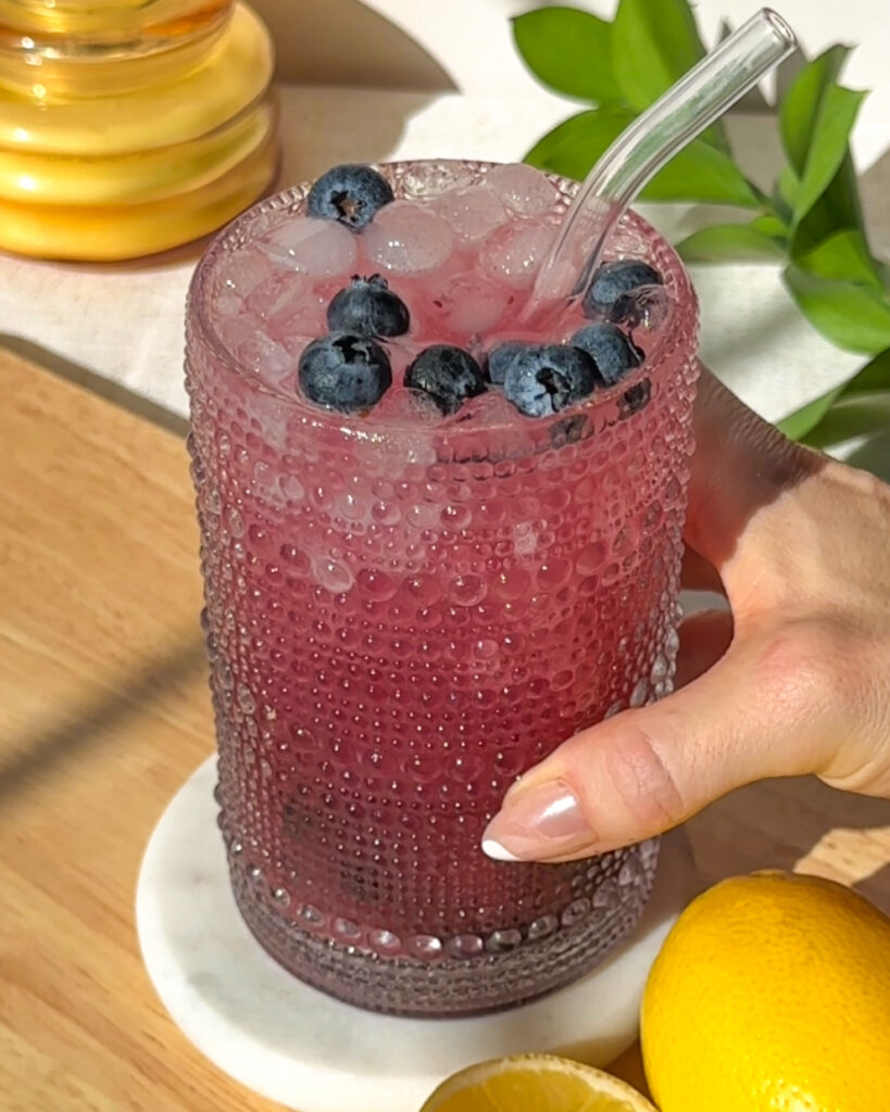 Hand holding a refreshing blueberry hydration drink topped with fresh blueberries and served over ice.