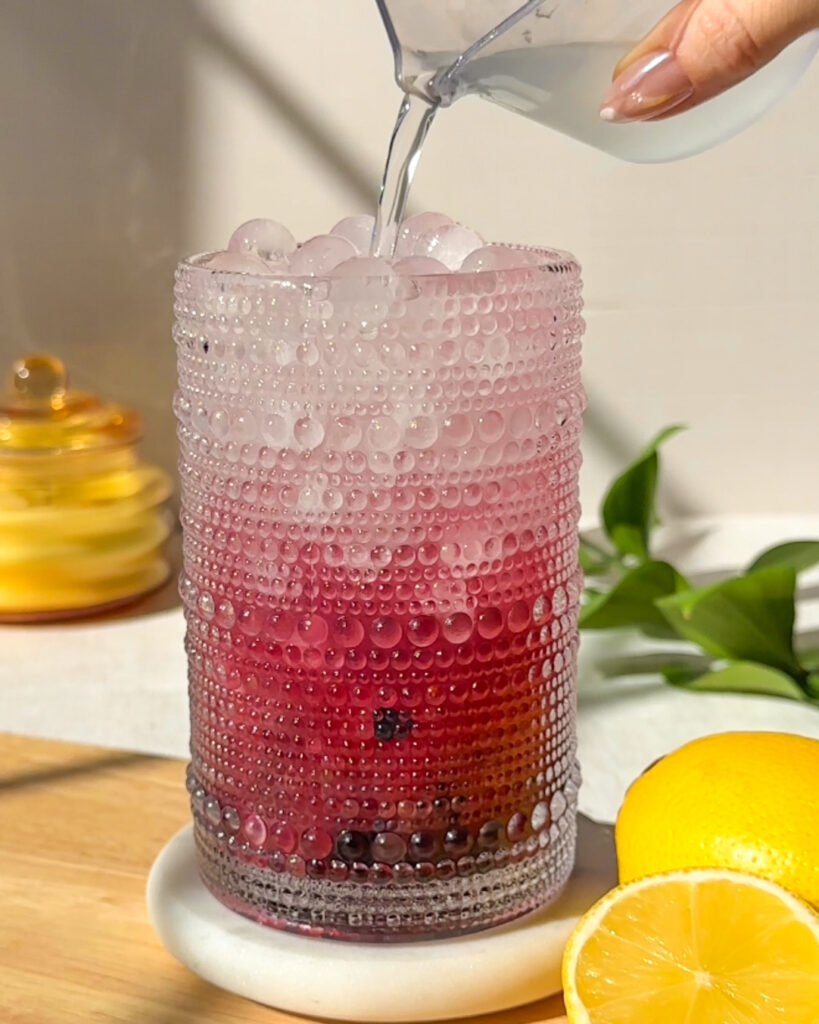 Pouring coconut water into a homemade blueberry hydration drink with fresh blueberries and ice.