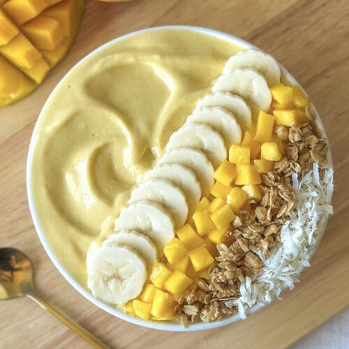 Thick mango smoothie bowl topped with banana slices, fresh mango, granola, and coconut on a wooden board with mangoes in the background