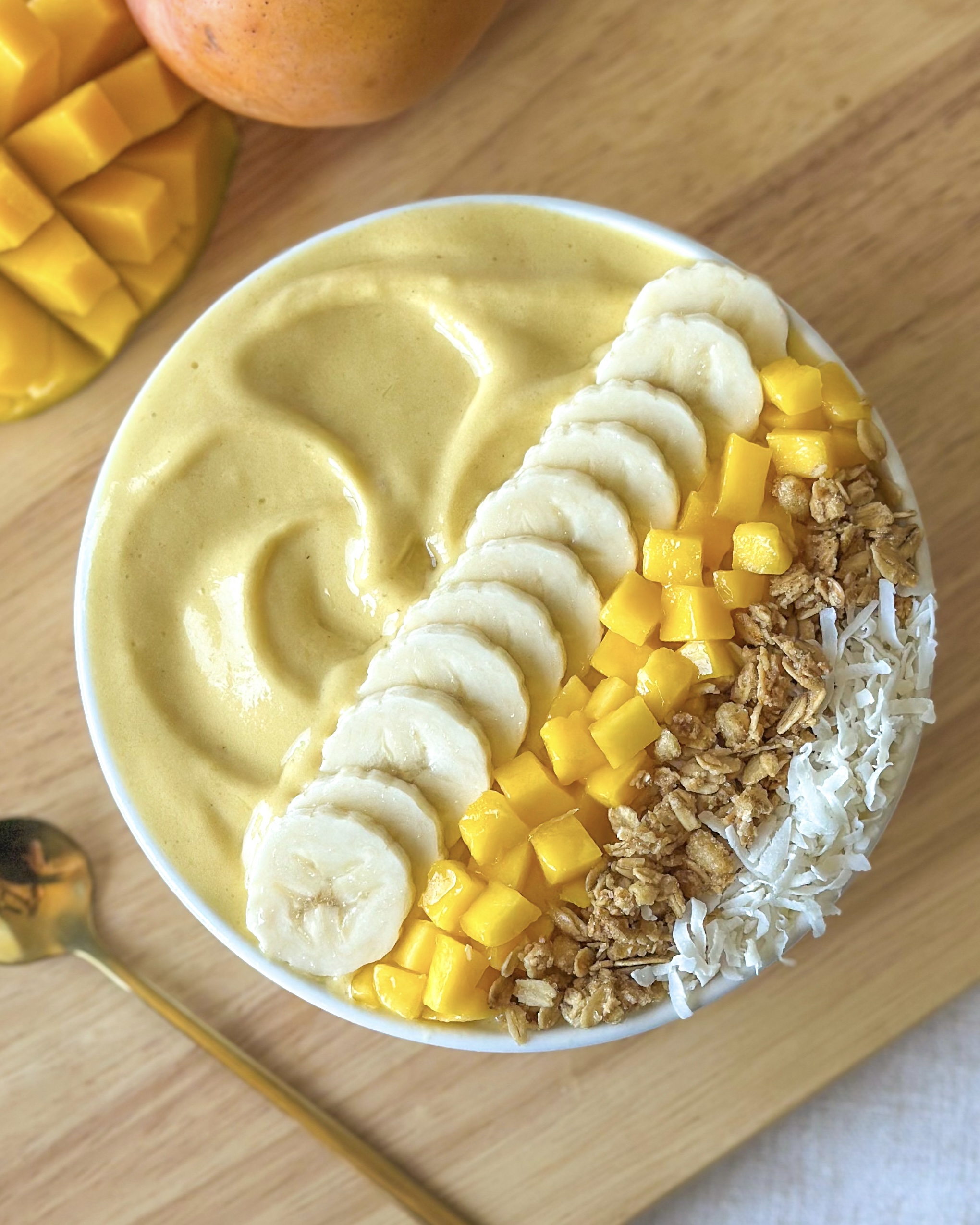 Thick mango smoothie bowl topped with banana slices, fresh mango, granola, and coconut on a wooden board with mangoes in the background