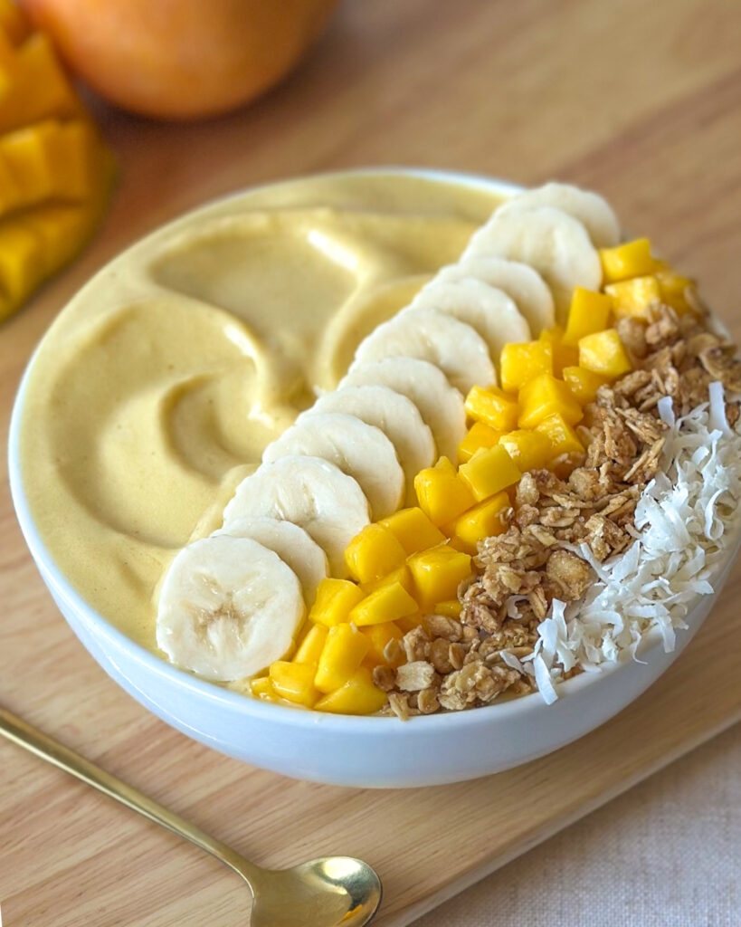 Front view of a mango smoothie bowl with a golden spoon and fresh mangoes in the background