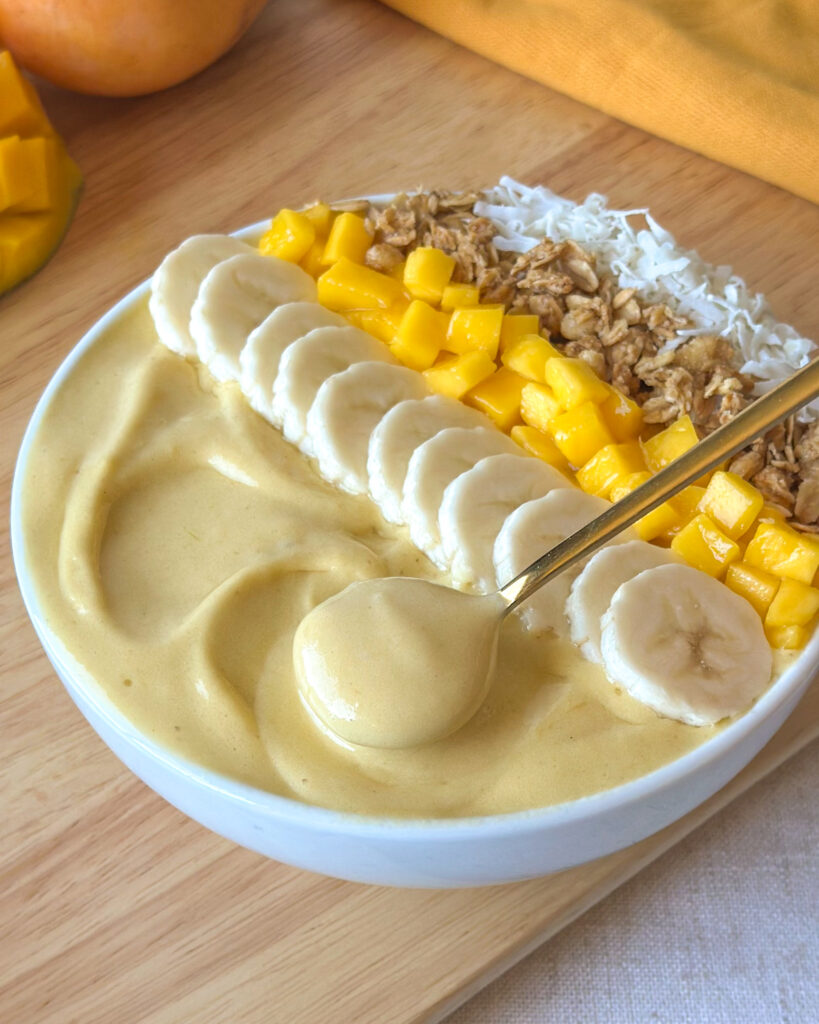 Overhead angled view of a mango smoothie bowl with a spoon lifting the thick smoothie texture