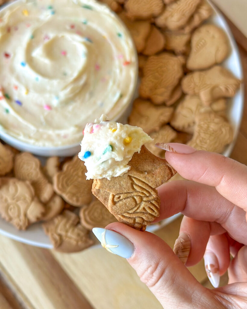 Hand holding an animal cracker topped with homemade Dunkaroo dip with the dip blurred in the background.