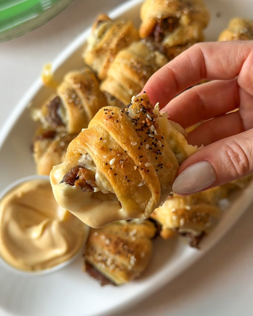 Hand holding a cheesy steak crescent roll bite dipped in sriracha mayonnaise sauce