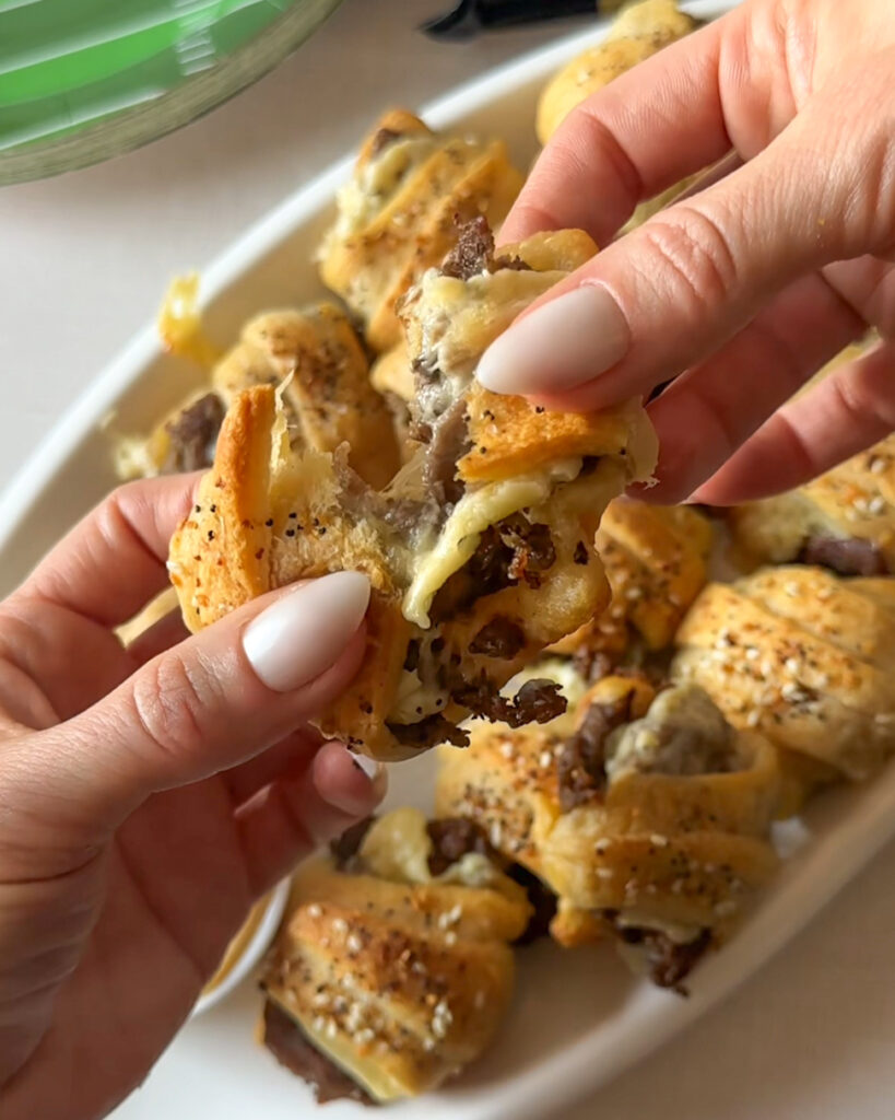 Hands pulling apart a cheesy steak crescent roll bite to reveal melted cheddar cheese and steak inside