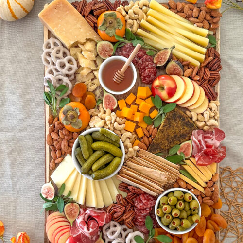 Overhead view of a fall charcuterie board filled with cheeses, cured meats, fruit, nuts, crackers, and honey with pumpkins and flowers in the background