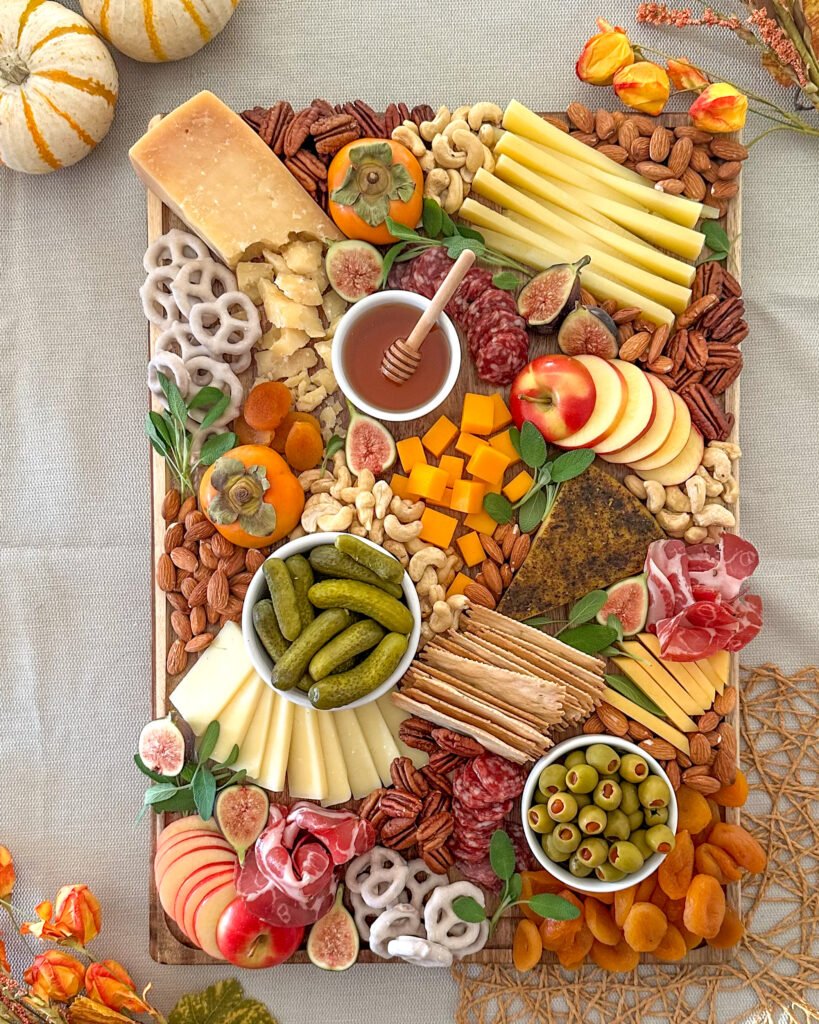 Overhead view of a fall charcuterie board filled with cheeses, cured meats, fruit, nuts, crackers, and honey with pumpkins and flowers in the background