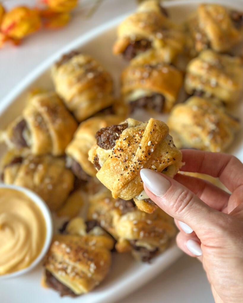 Hands holding a cheesy steak crescent roll bite with more finished bites in the background