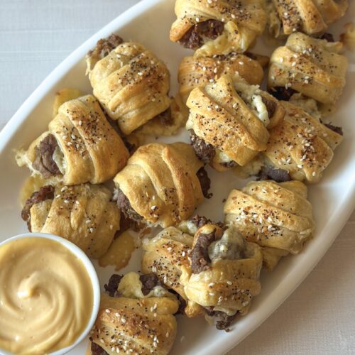 Overhead view of cheesy steak crescent roll bites served on an oval platter with sriracha mayonnaise dipping sauce