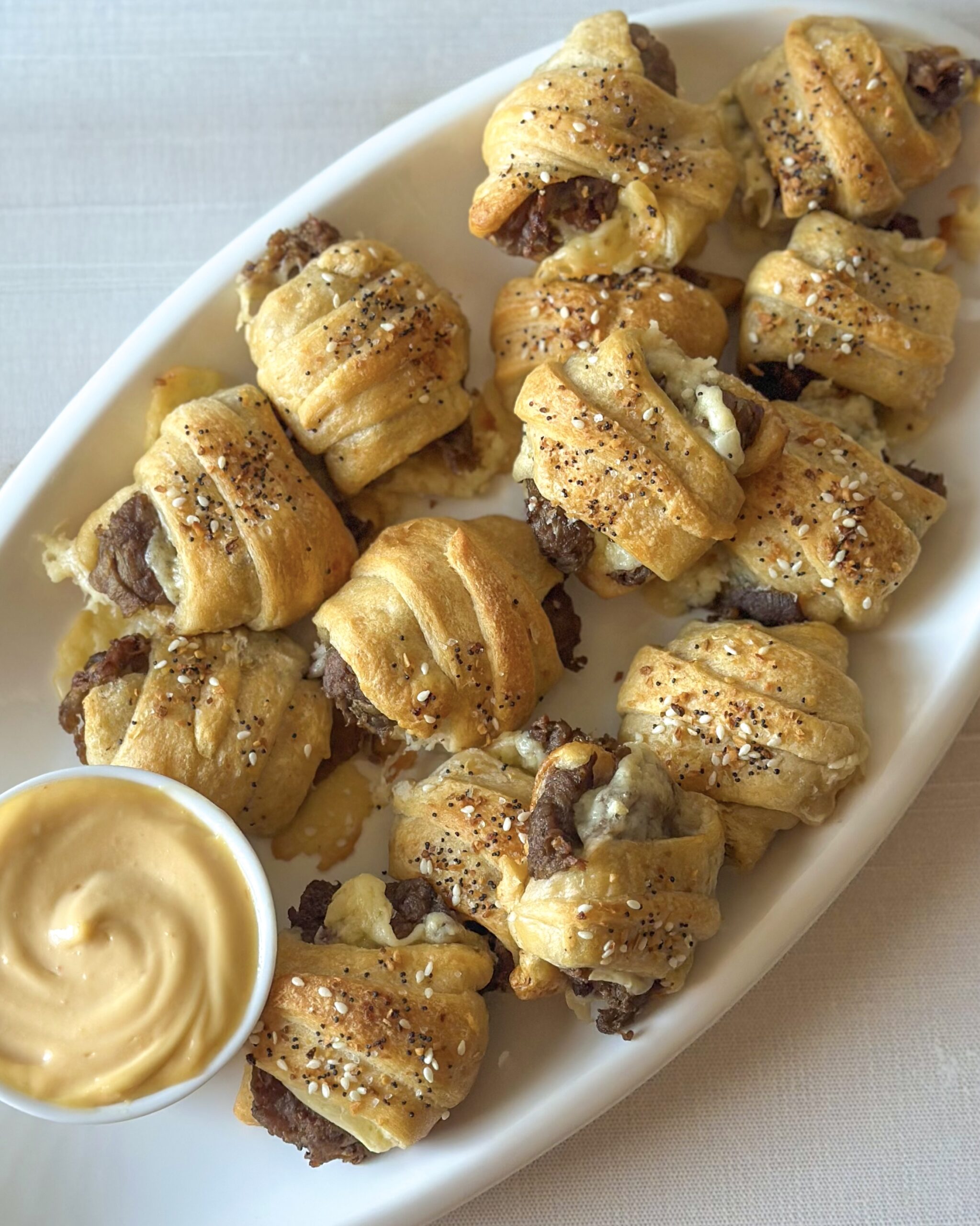 Overhead view of cheesy steak crescent roll bites served on an oval platter with sriracha mayonnaise dipping sauce